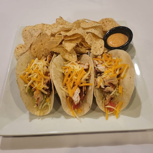 Three chicken tacos with shredded cheese, tortilla chips, and a side of dipping sauce on a white plate.