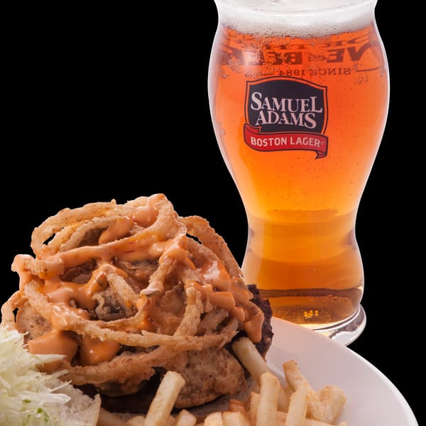 A burger topped with onion rings and sauce, with fries and a Samuel Adams Boston Lager.