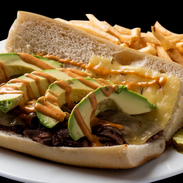 A steak sandwich with melted cheese, avocado slices, and sauce, served with fries and pickles.