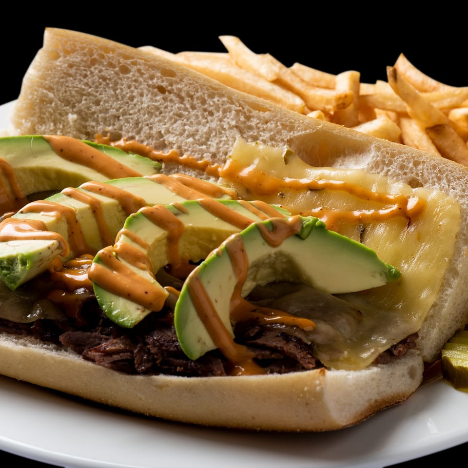 A steak sandwich with melted cheese, avocado slices, and sauce, served with fries and pickles.