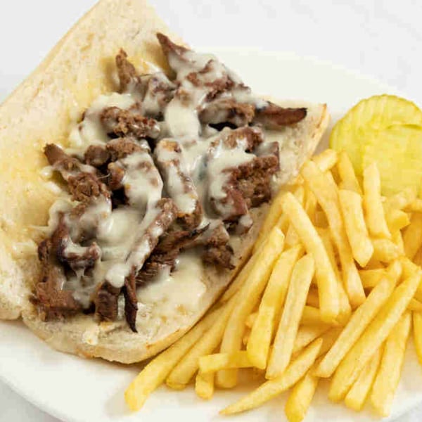 Cheesesteak sandwich with melted cheese and a side of golden french fries and a pickle.