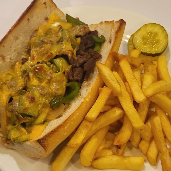 Cheesesteak sandwich with green peppers, onions, and melted cheese, served with fries and pickles.