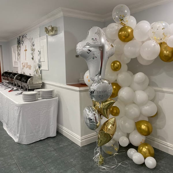 A buffet table with chafing dishes and stacked plates, next to a white and gold balloon arch.