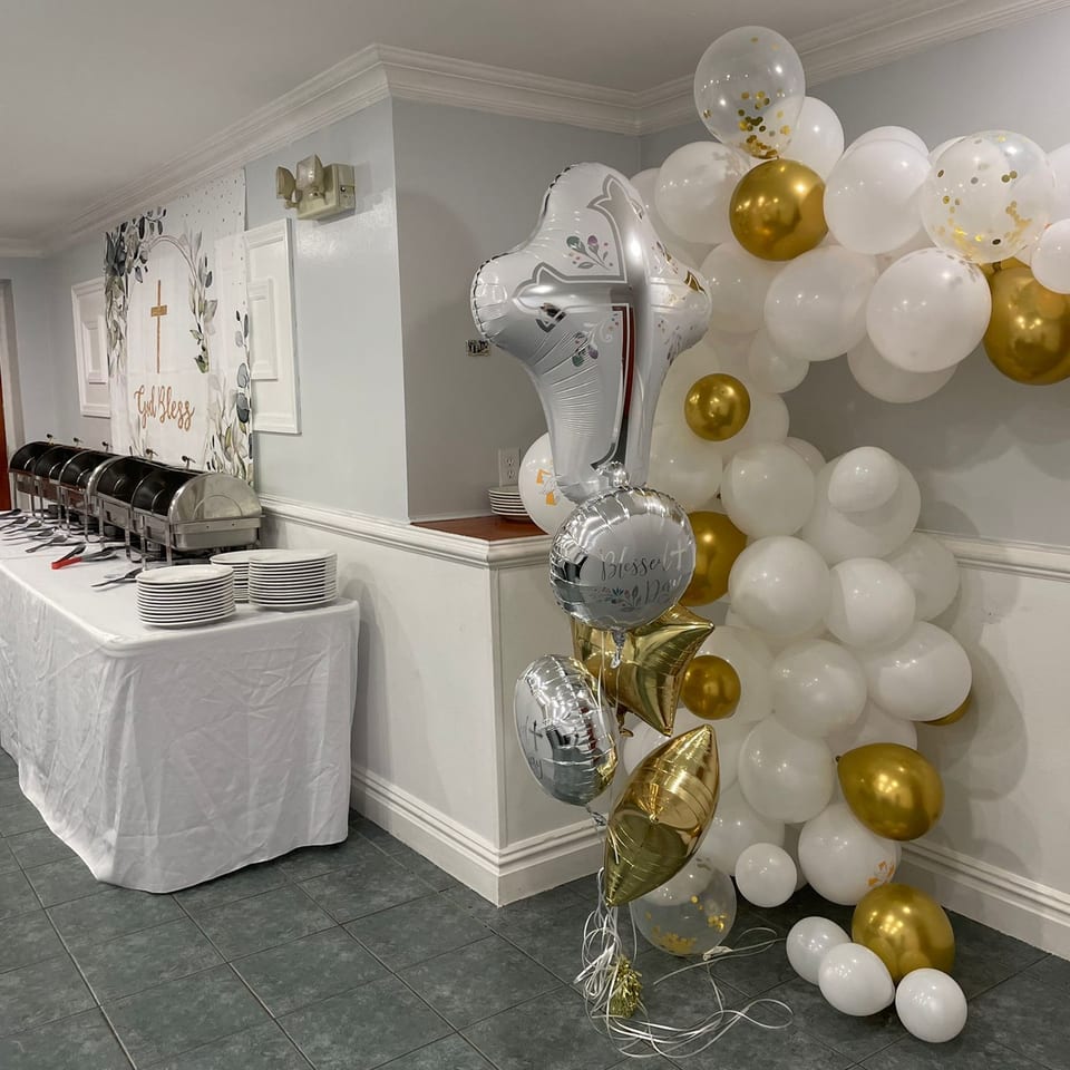 A buffet table with chafing dishes and stacked plates, next to a white and gold balloon arch.
