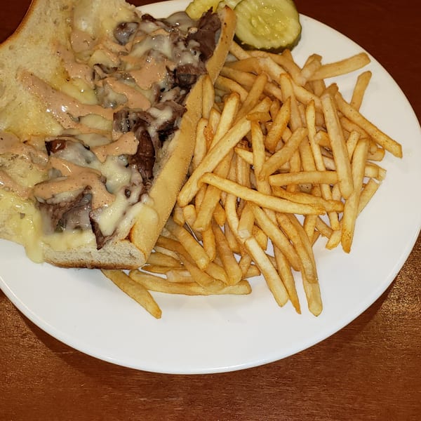 Cheesesteak sandwich with melted cheese and sauce, served with french fries and pickles on a white plate.