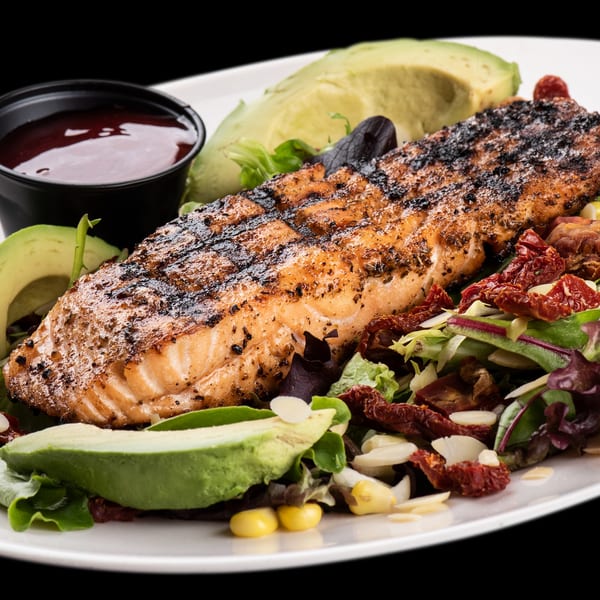 Grilled salmon fillet with avocado slices, mixed greens, sun-dried tomatoes, and a side of sauce.