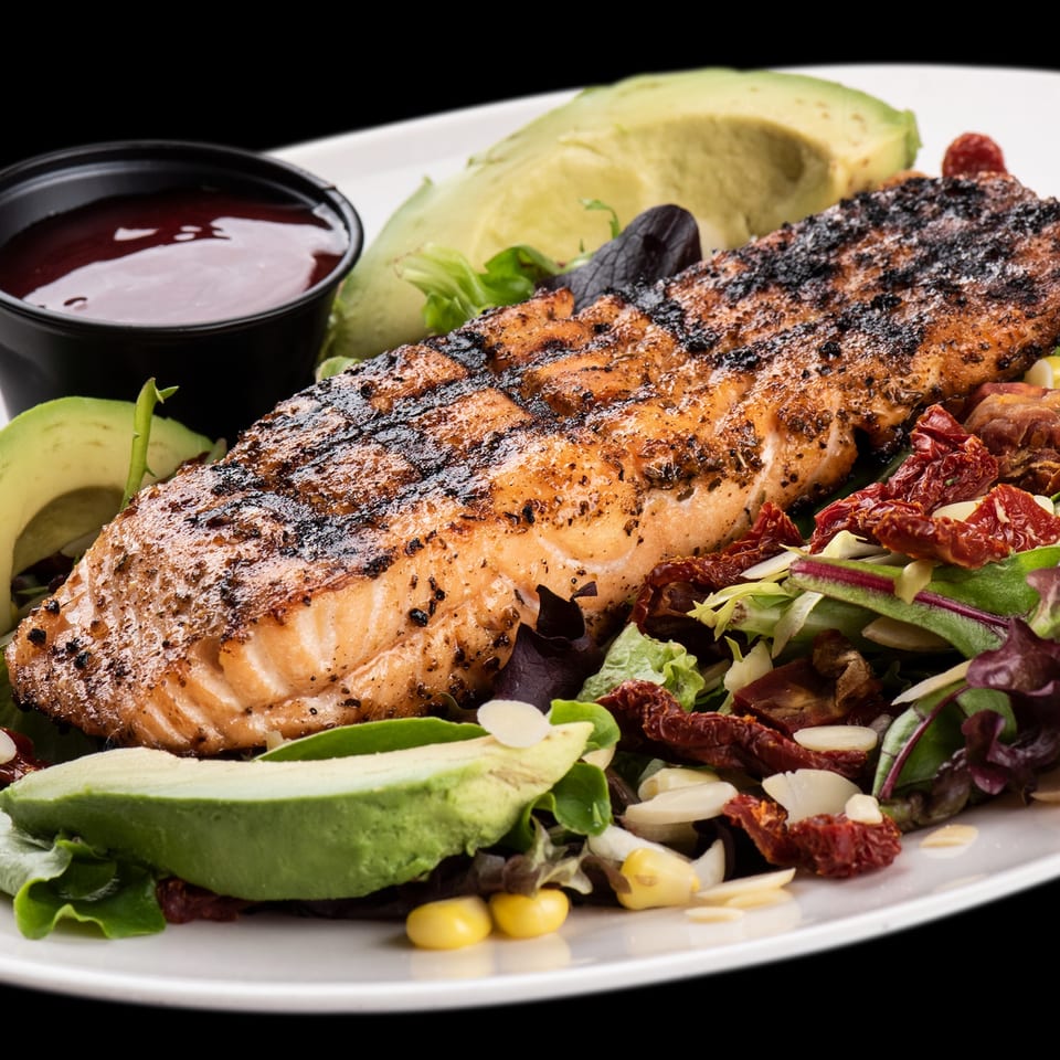 Grilled salmon fillet with avocado slices, mixed greens, sun-dried tomatoes, and a side of sauce.