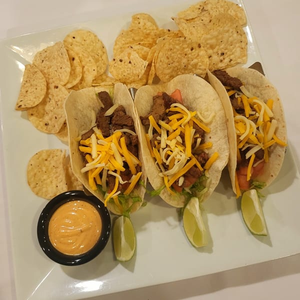 Three steak tacos with cheese, lettuce, and tomatoes, served with tortilla chips, lime wedges, and sauce.