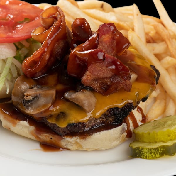A close-up of a Town Pub burger with bacon, mushrooms, cheese, and BBQ sauce, served with fries and pickles.